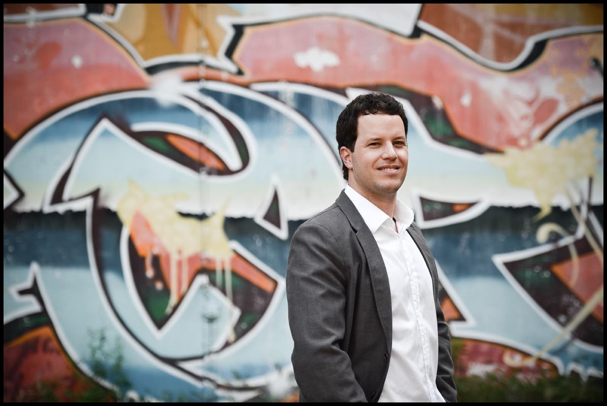 Ramón smiles wearing a dark gray suit and white button-down shirt with the top button open. He's standing in front of a wall of graffiti letters in blue and burnt orange